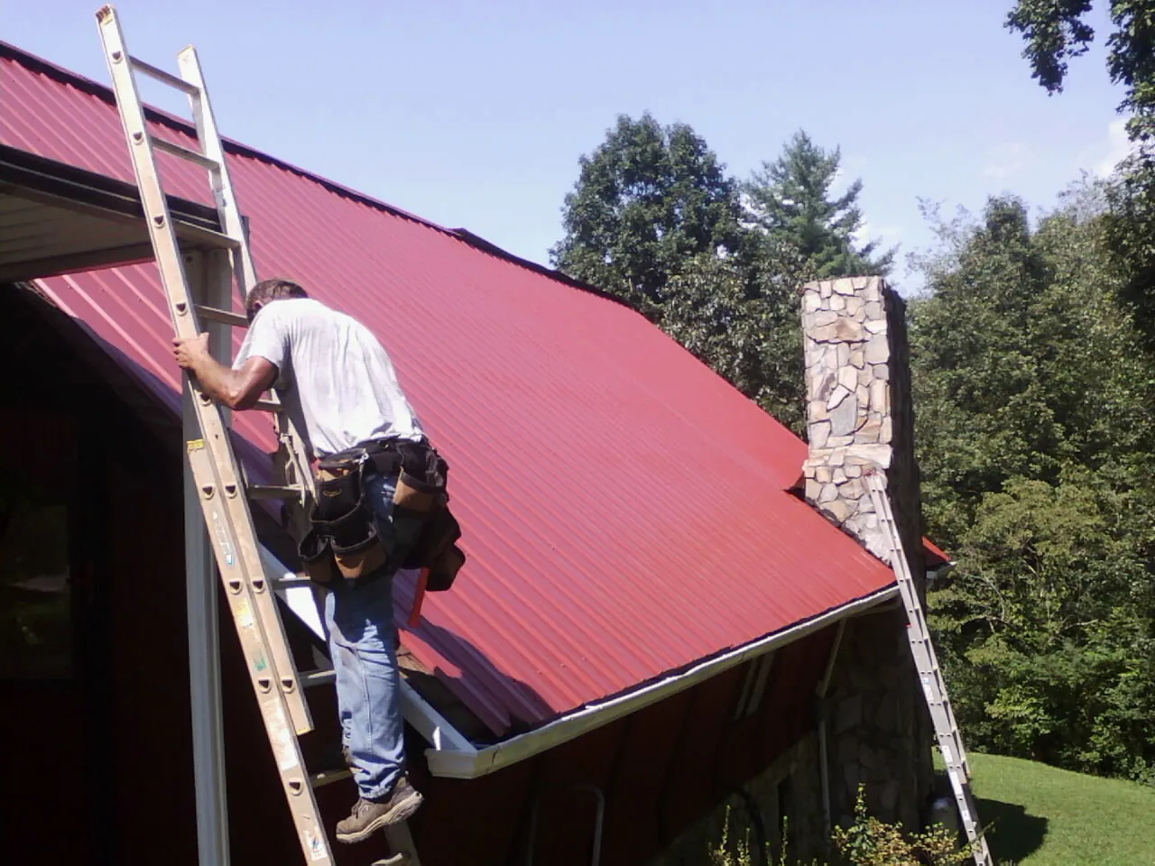 Roofer on a ladder beside a red metal roof and stone chimney — Soffit & Fascia Repair by Roofing Brothers in Kremlin